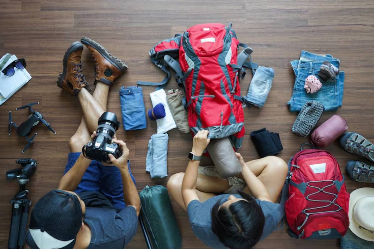 Aerial view of two people packing a red backpack with clothing and gear, while one holds a camera, preparing for an outdoor adventure.