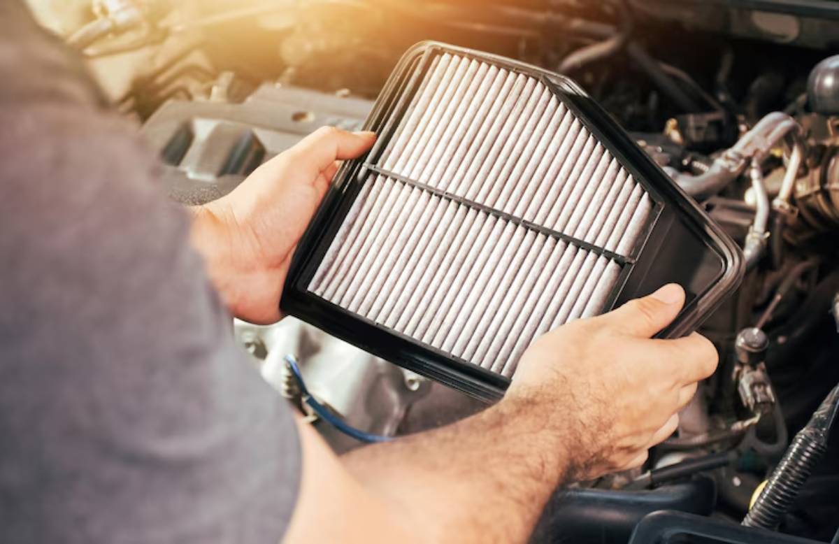 A person holds a clean air filter above an open car engine, preparing for maintenance or replacement, with sunlight filtering in.