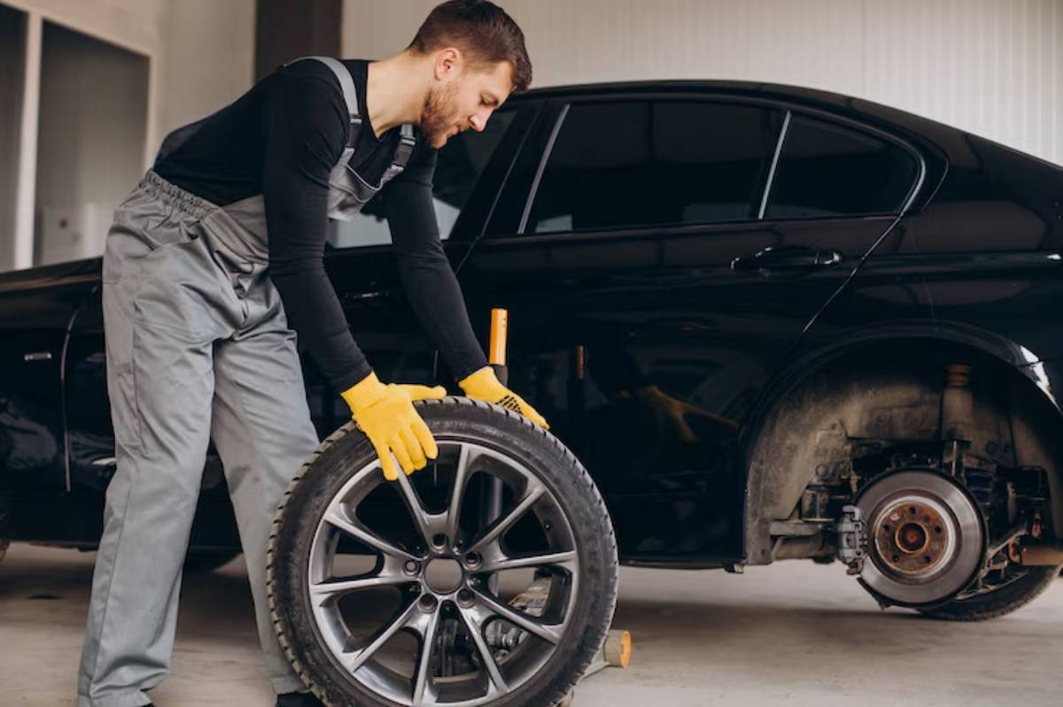 A mechanic in gray overalls and yellow gloves lifts a car wheel in a garage, beside a black vehicle with a missing tire.