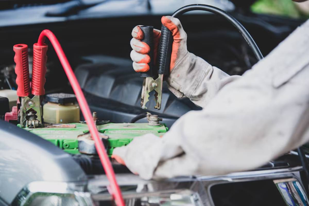 A person in a work glove holds a jumper cable clamp above a green car battery, preparing to jump-start the vehicle.