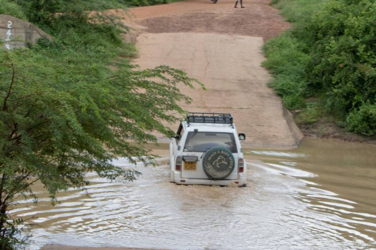 A white SUV drives through a flooded dirt road, surrounded by lush greenery, while a person walks in the distance.