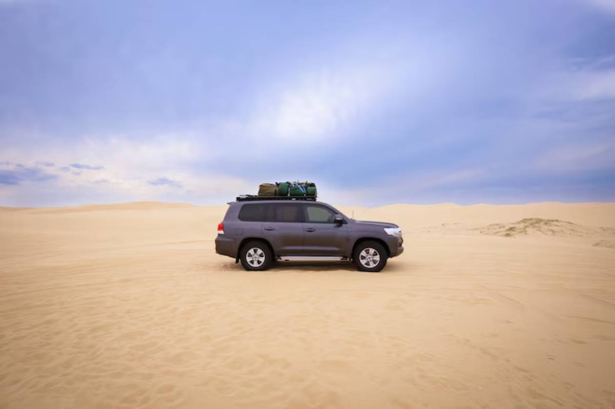 A grey SUV parked on sandy dunes, with camping gear strapped on the roof, against a cloudy sky.