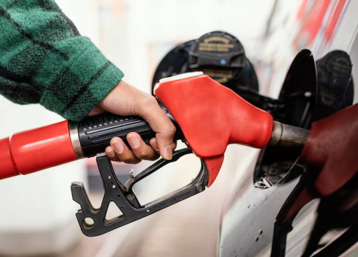 A hand holding a red gas pump nozzle, inserting it into a car's fuel tank at a gas station, wearing a green plaid shirt.