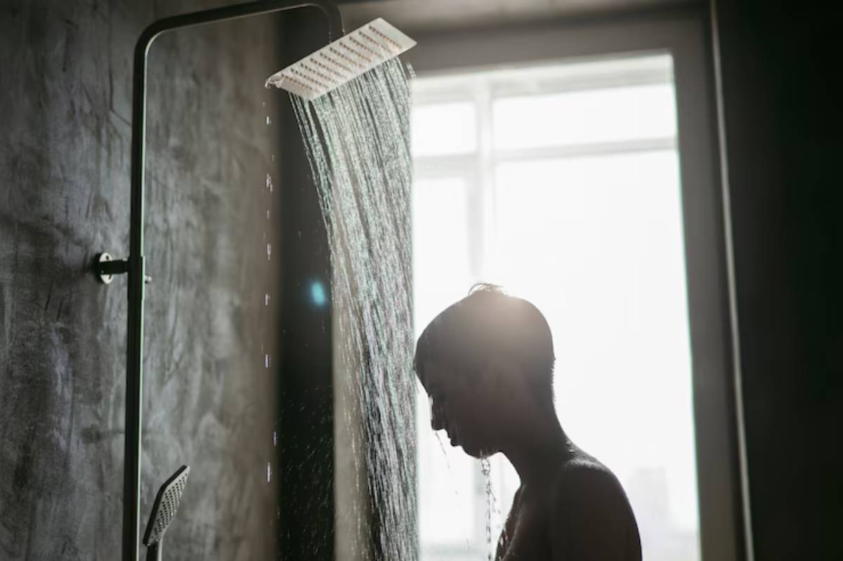 A silhouette of a person stands under a modern showerhead, with water cascading down against a soft, illuminated background.