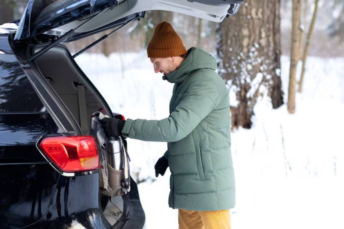 A person in a green puffer jacket and brown hat is reaching into the trunk of a black SUV parked in a snowy landscape.