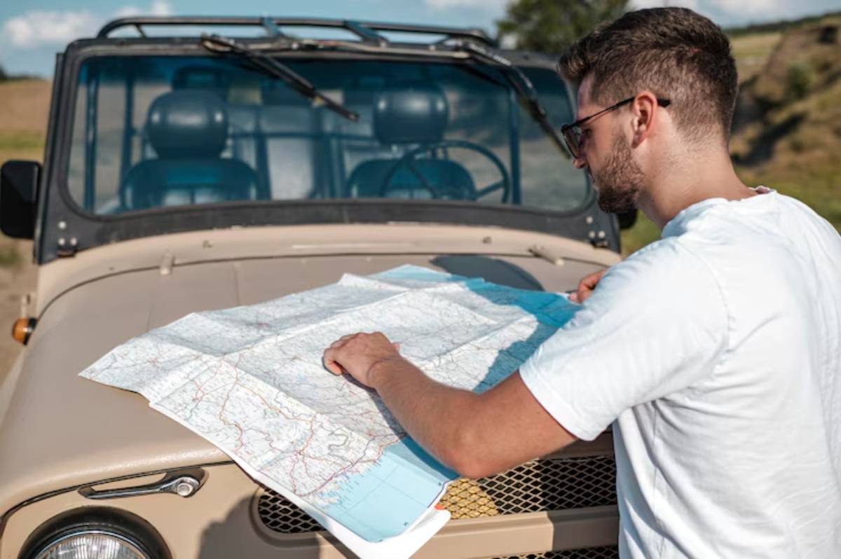 A person studies a detailed map, with a vintage jeep parked in the background under a clear blue sky and open countryside.