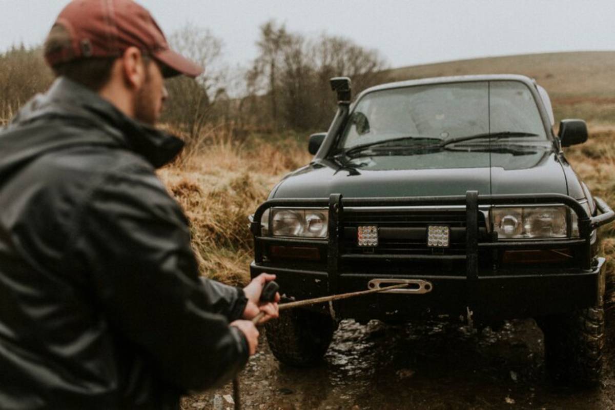 A man in a black jacket and cap prepares a rope in front of a rugged black off-road vehicle on a muddy terrain.