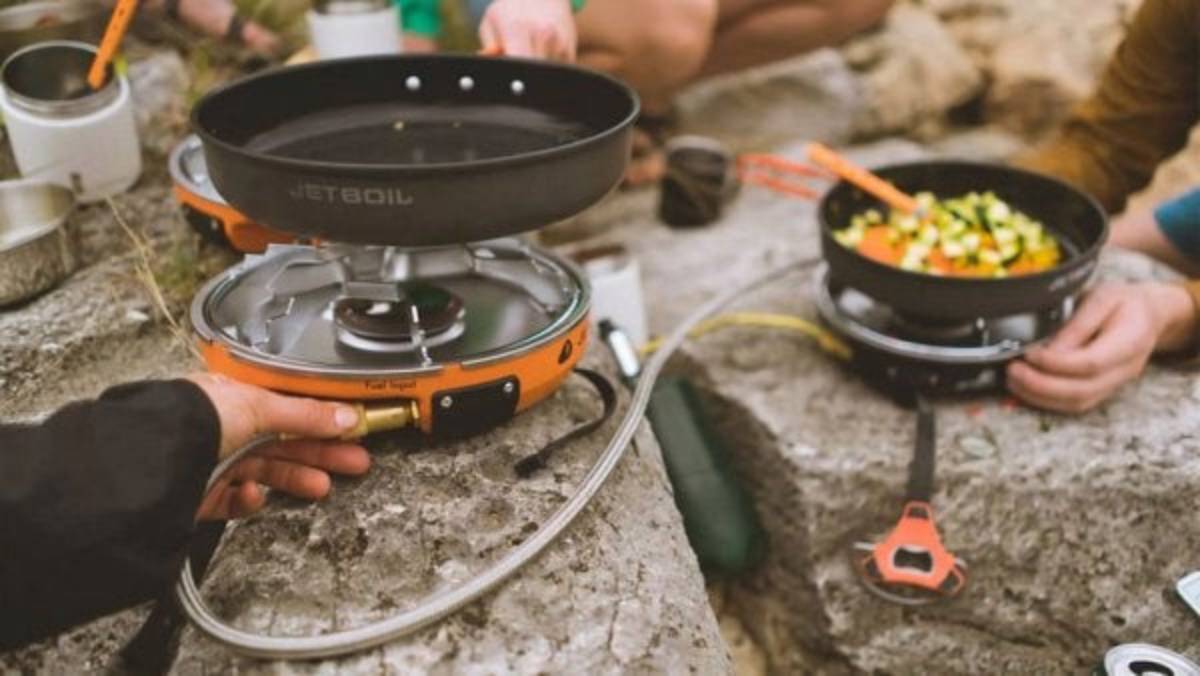 People cooking outdoors on rocky terrain using Jetboil Genesis Basecamp propane stoves, with one pan empty and the other filled with chopped vegetables being stirred with tongs