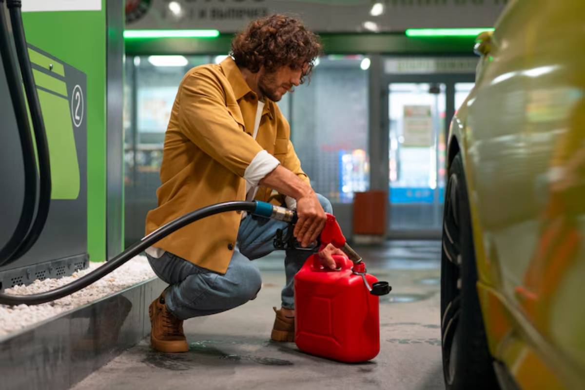 A person kneels at a gas station, filling a red canister with fuel from a pump, with a yellow car parked nearby.