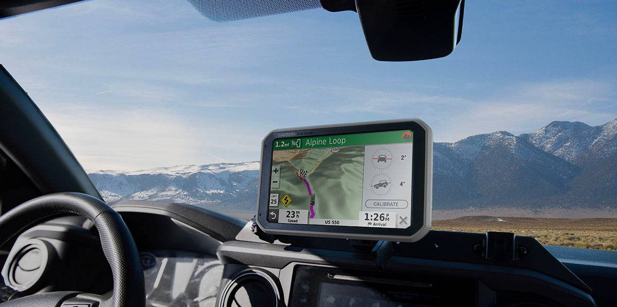 A GPS device displays a route through mountainous terrain alongside a car's steering wheel, with snow-capped mountains in the background.