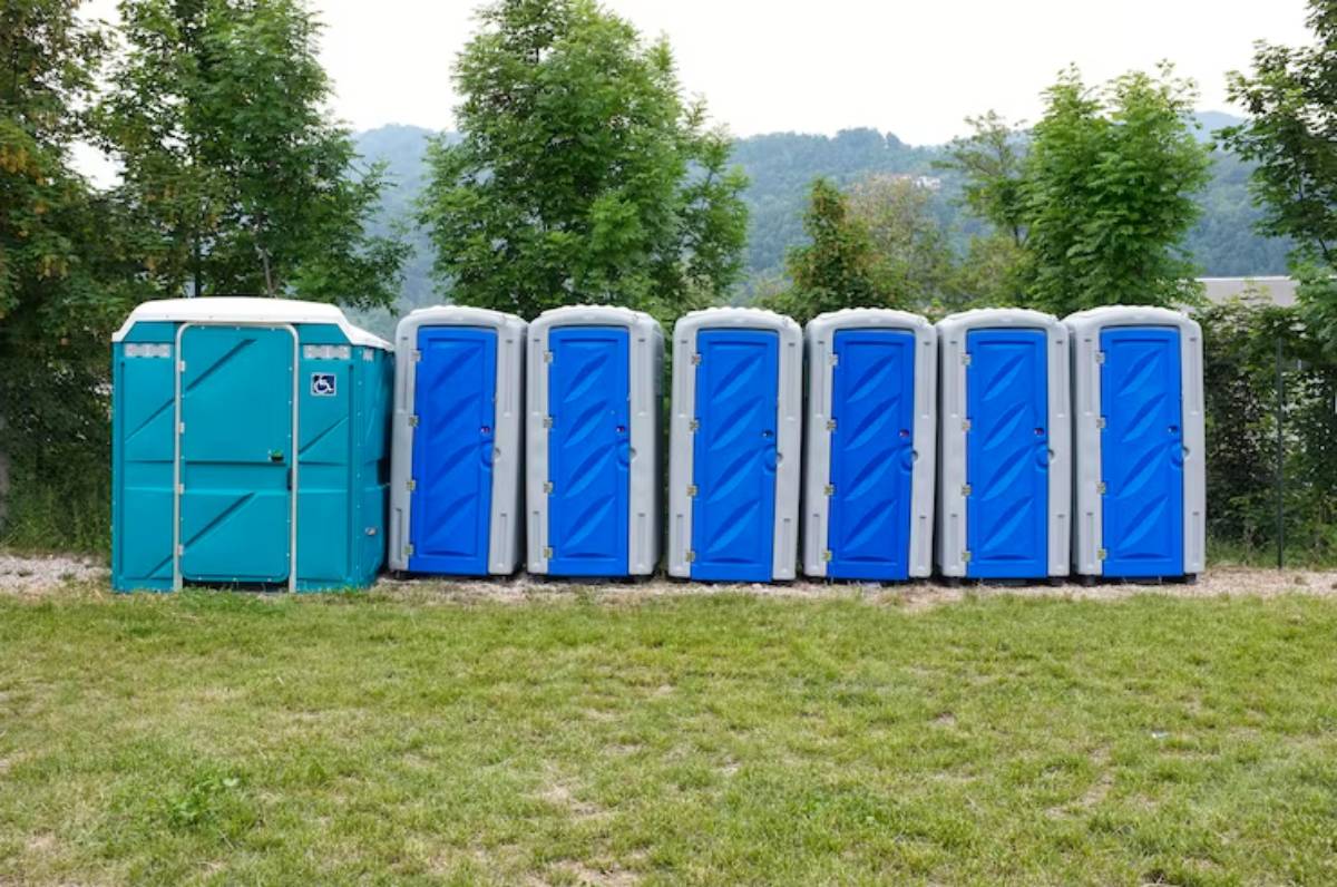 A row of eight portable toilets, including one accessible unit, situated on grass with trees in the background under a cloudy sky.