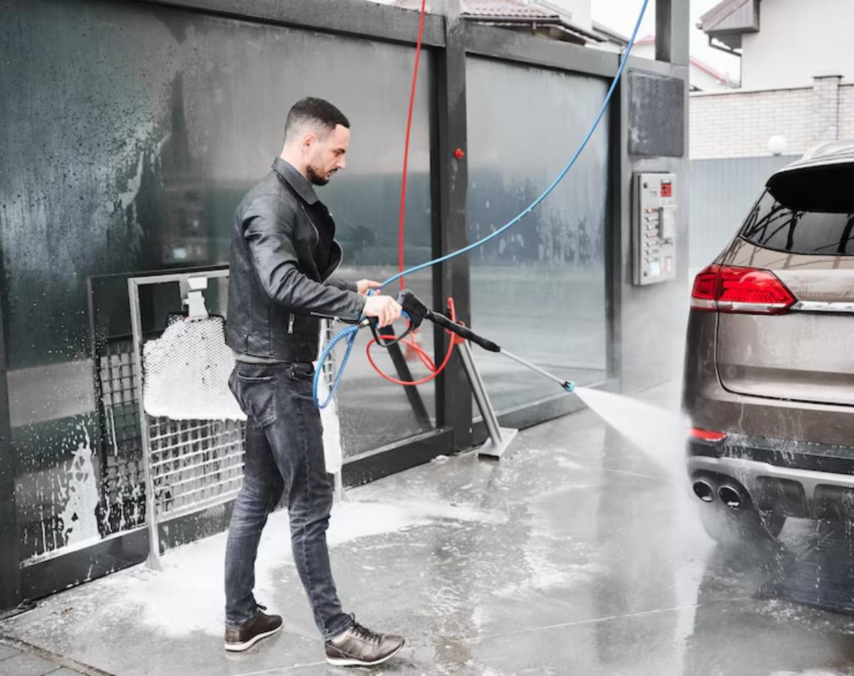 A person washes a brown SUV with a pressure washer at a car wash, surrounded by soap suds and water spray.