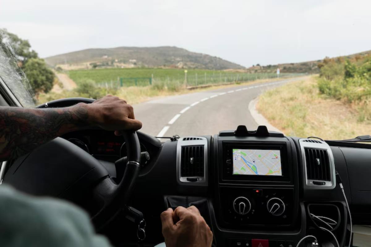 A driver's hand grips the steering wheel of a van with a GPS screen, navigating a winding rural road lined with vineyards.