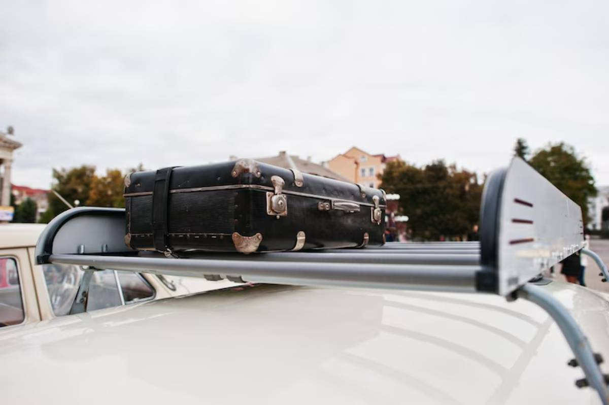 A classic black suitcase rests on the roof rack of a vintage car, with a cloudy sky and colorful buildings in the background.