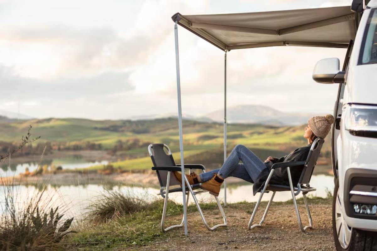 A person relaxes under an awning beside a van, overlooking a serene lake and lush greenery in the background.