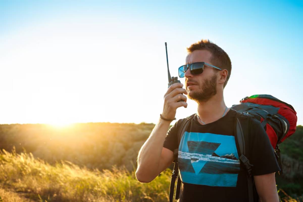 A backpacker holding a walkie-talkie against a sunset backdrop, surrounded by grassy terrain and clear blue skies.