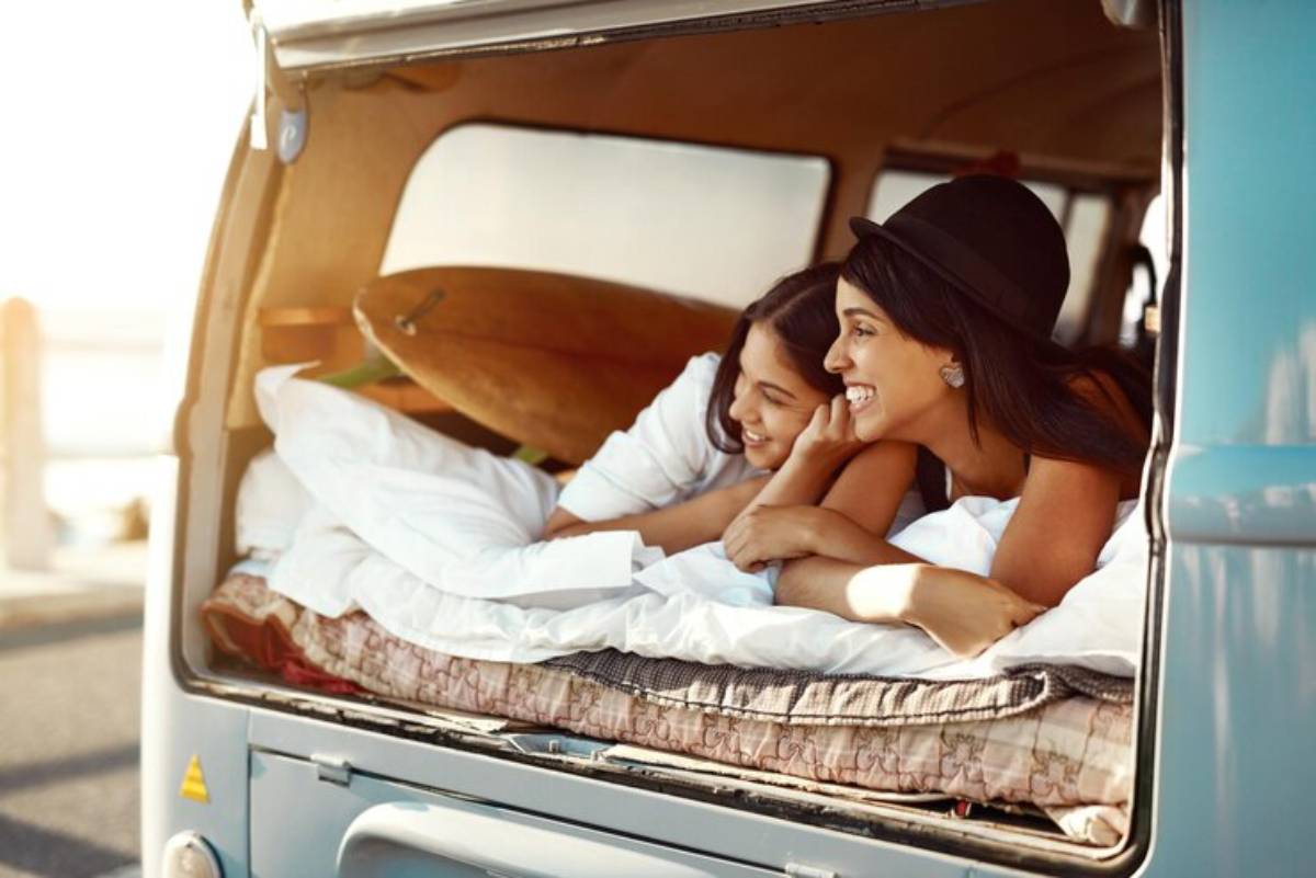 A couple relaxes in a cozy van, surrounded by white bedding and a surfboard, enjoying the warm sunset light.