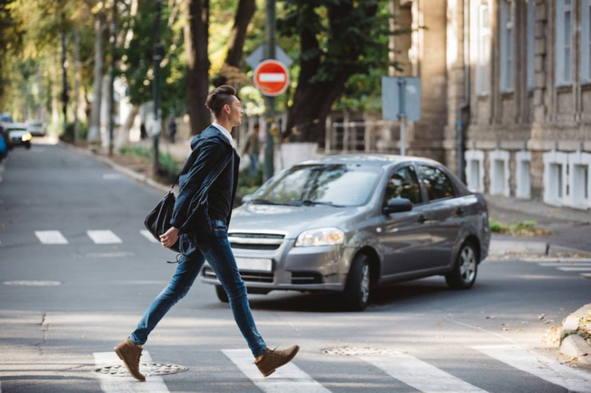 A young man in casual attire walks across a crosswalk, with a car approaching in a tree-lined street.