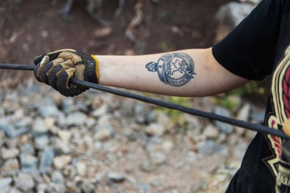 Person wearing protective gloves while handling a steel cable outdoors, with a visible tattoo on their forearm and rocky terrain in the background