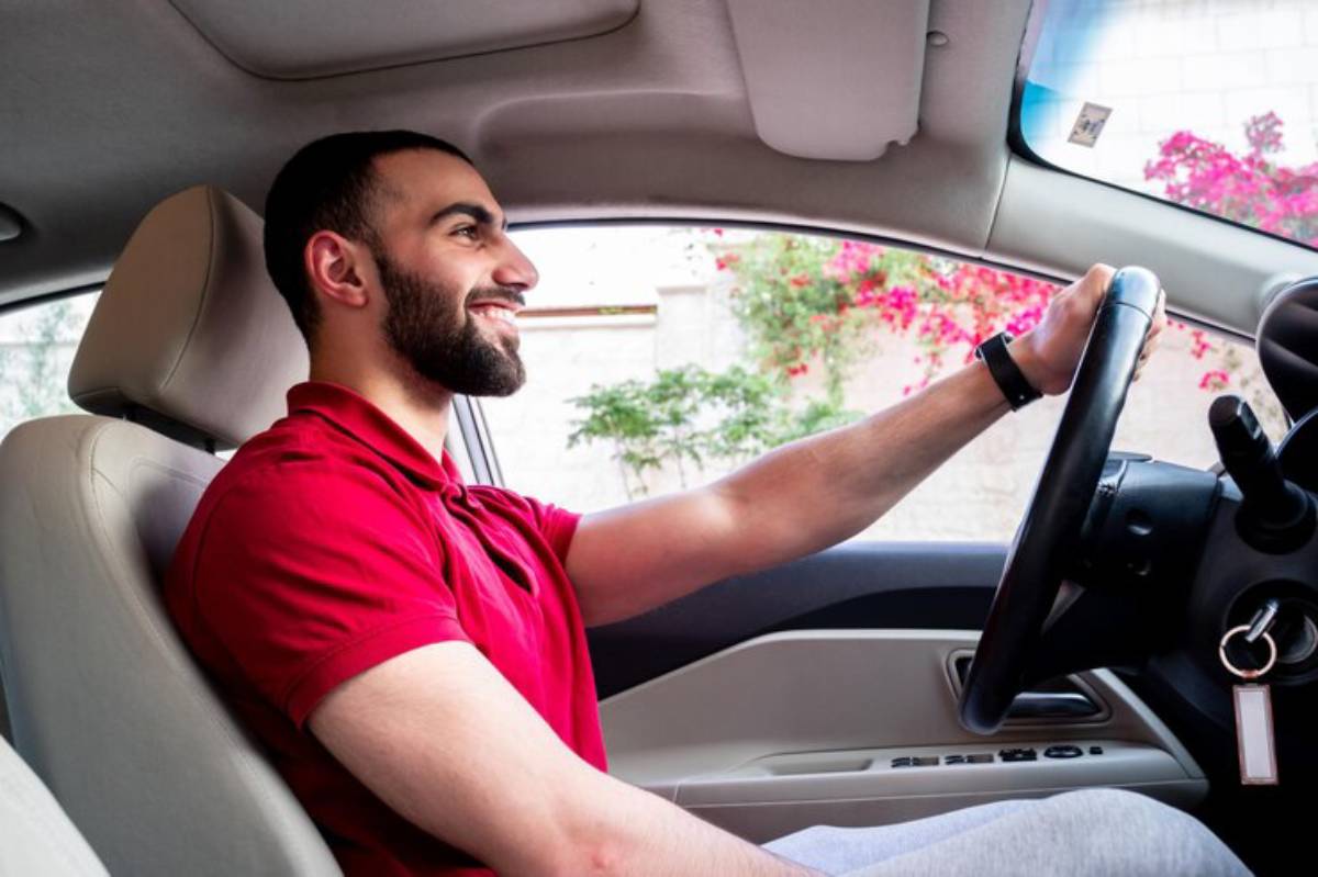 A person in a red polo shirt is seated in a car, gripping the steering wheel, with colorful flowers visible outside the window.
