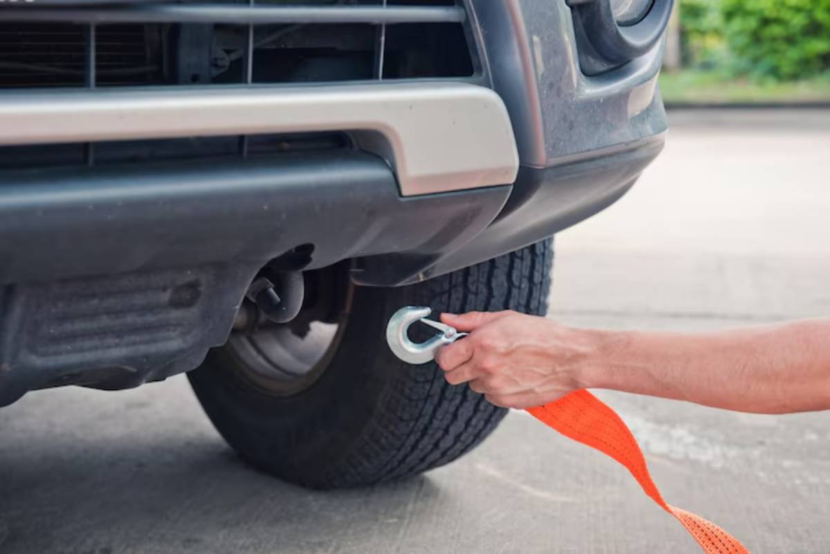 A person attaches a metal hook to the rear of a vehicle using an orange towing strap for assistance or recovery.