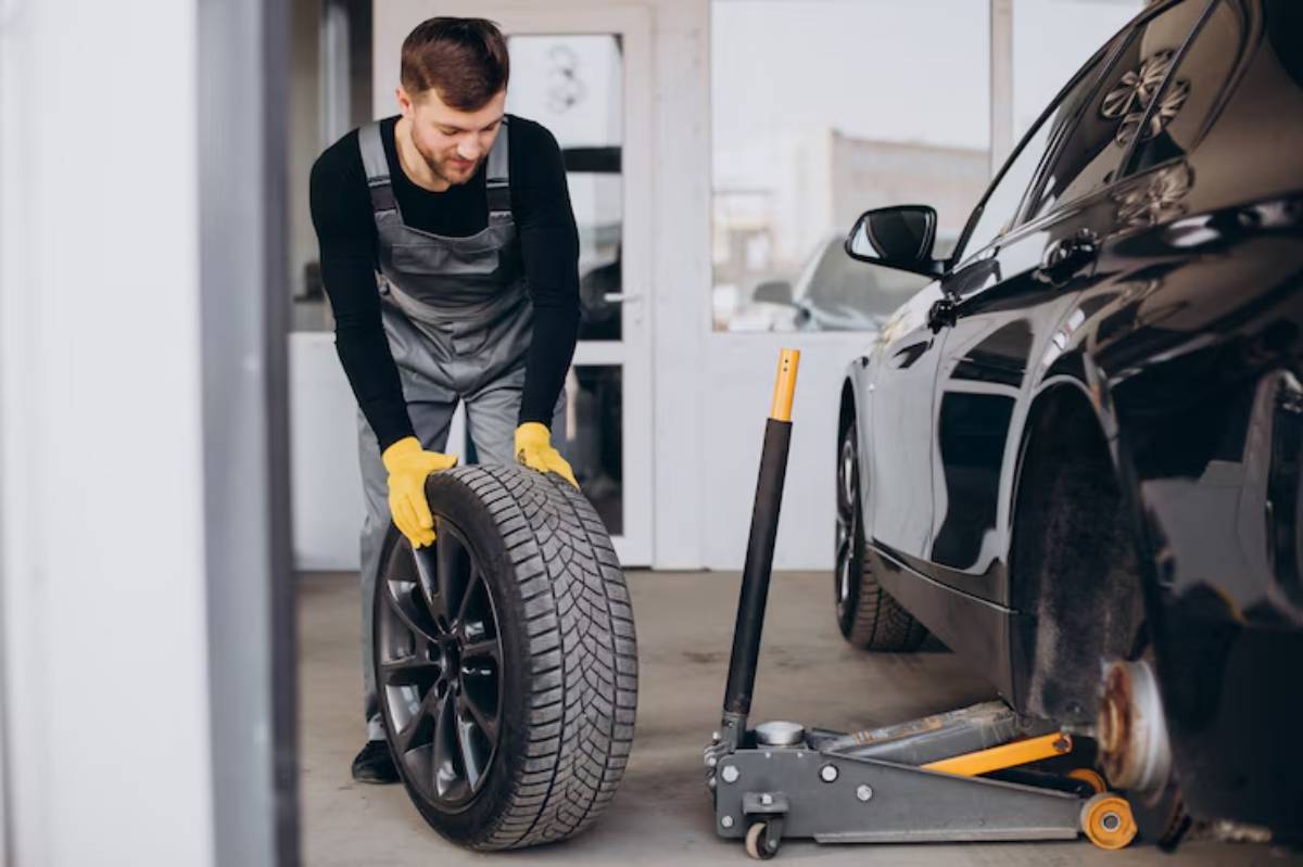 A mechanic in gloves is lifting a tire in a garage, with a black car and a jack visible in the background.