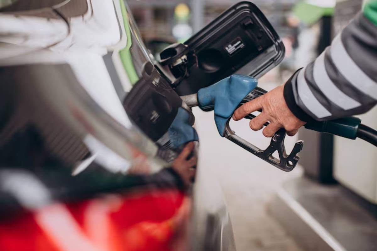 A person holds a fuel pump nozzle, preparing to refuel a vehicle at a gas station. The car's rear is visible, showcasing the shiny surface.