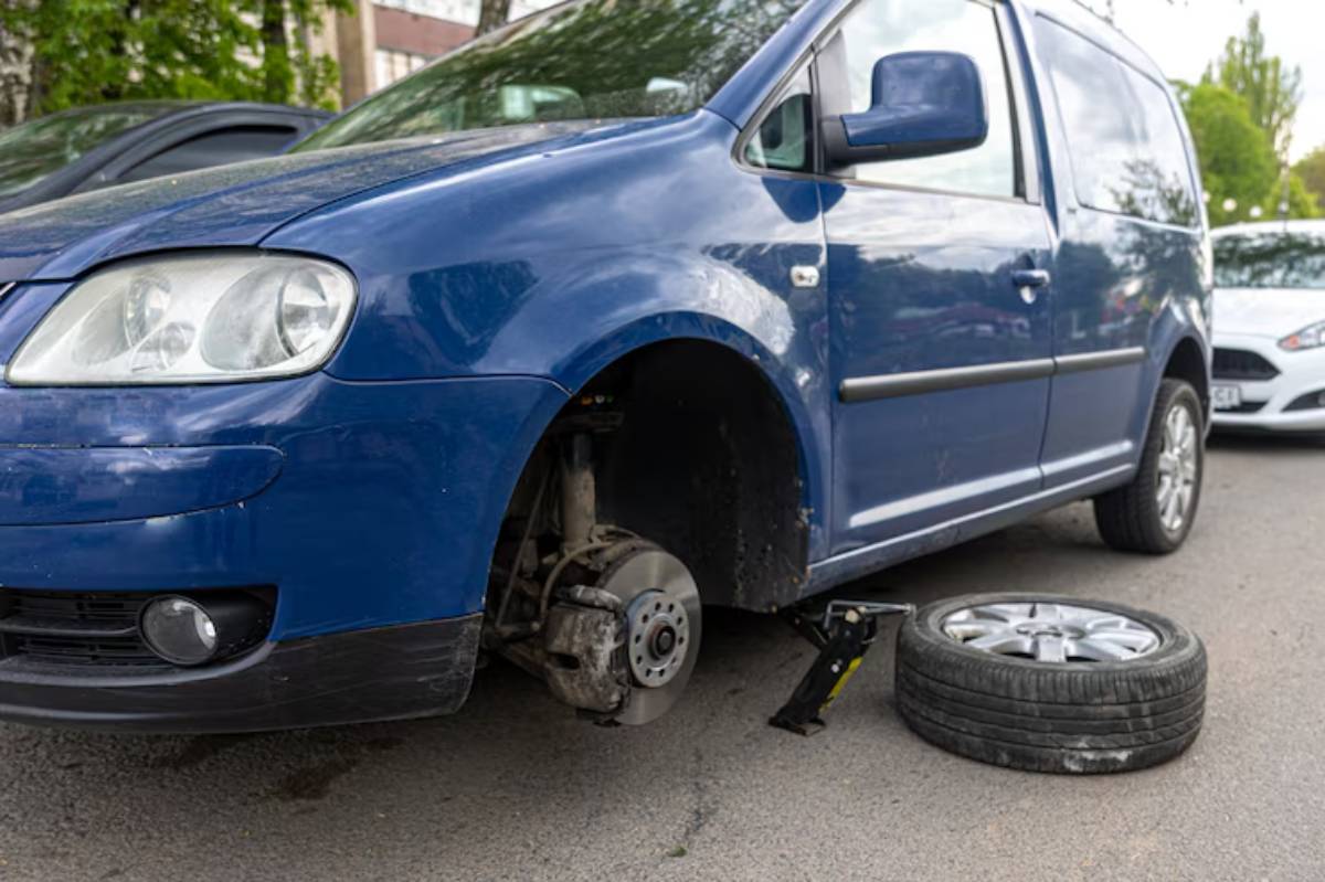 A blue van with one wheel removed, propped up on a jack, next to its detached tire lying on the ground.