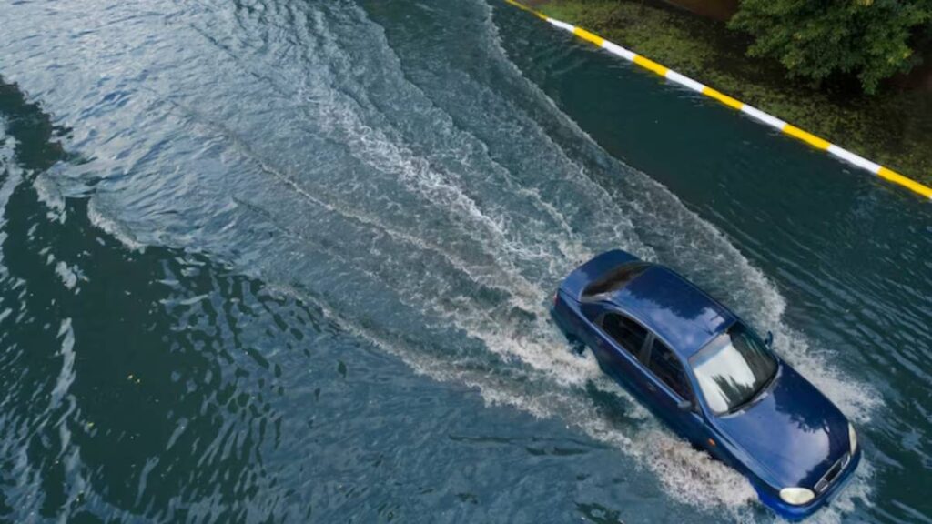 A blue car drives through a flooded street, creating ripples in the water as it moves past a yellow-striped curb.