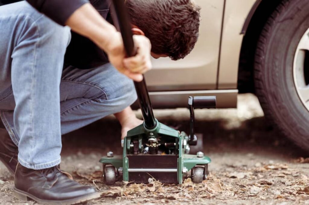 A man uses a green hydraulic jack to lift a vehicle on a gravel surface, preparing for tire maintenance or repair.