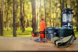 Camping equipment set on a wooden table in a forest, including a gas lantern, fuel bottle, portable stove, metal cookware