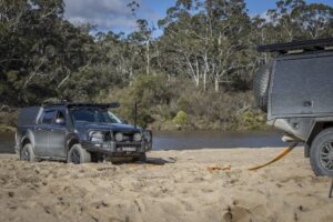 A rugged 4x4 vehicle is partially buried in sand near a river, connected to an off-road trailer with recovery straps amidst lush greenery.