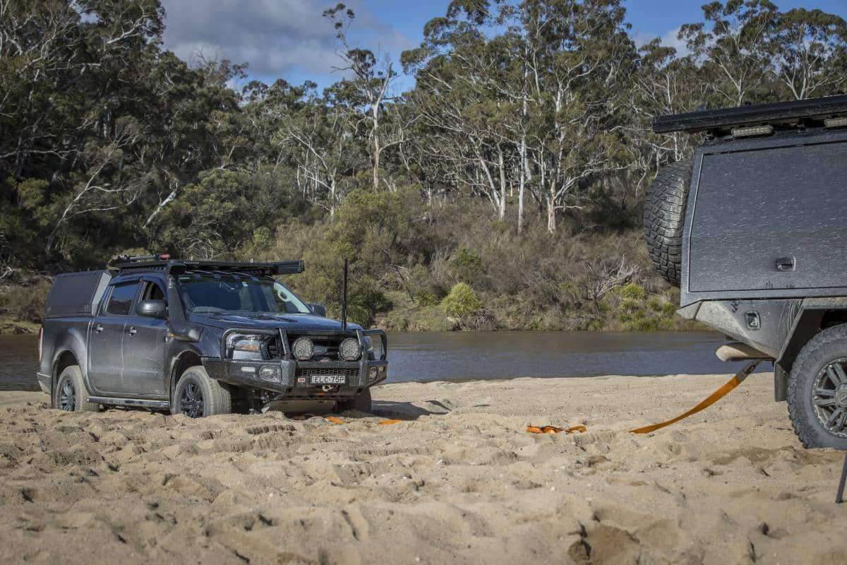 A rugged 4x4 vehicle is partially buried in sand near a river, connected to an off-road trailer with recovery straps amidst lush greenery.