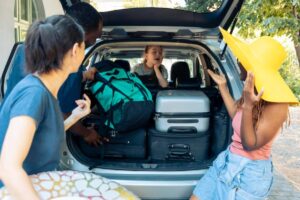 A group of friends are loading luggage into a car trunk, with colorful bags and a bright yellow hat enhancing the vibrant atmosphere.