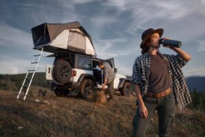 A woman in a checkered shirt drinks from a stainless steel bottle near a Jeep with a rooftop tent in a scenic outdoor setting.