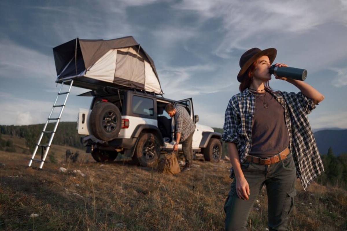 A woman in a checkered shirt drinks from a stainless steel bottle near a Jeep with a rooftop tent in a scenic outdoor setting.