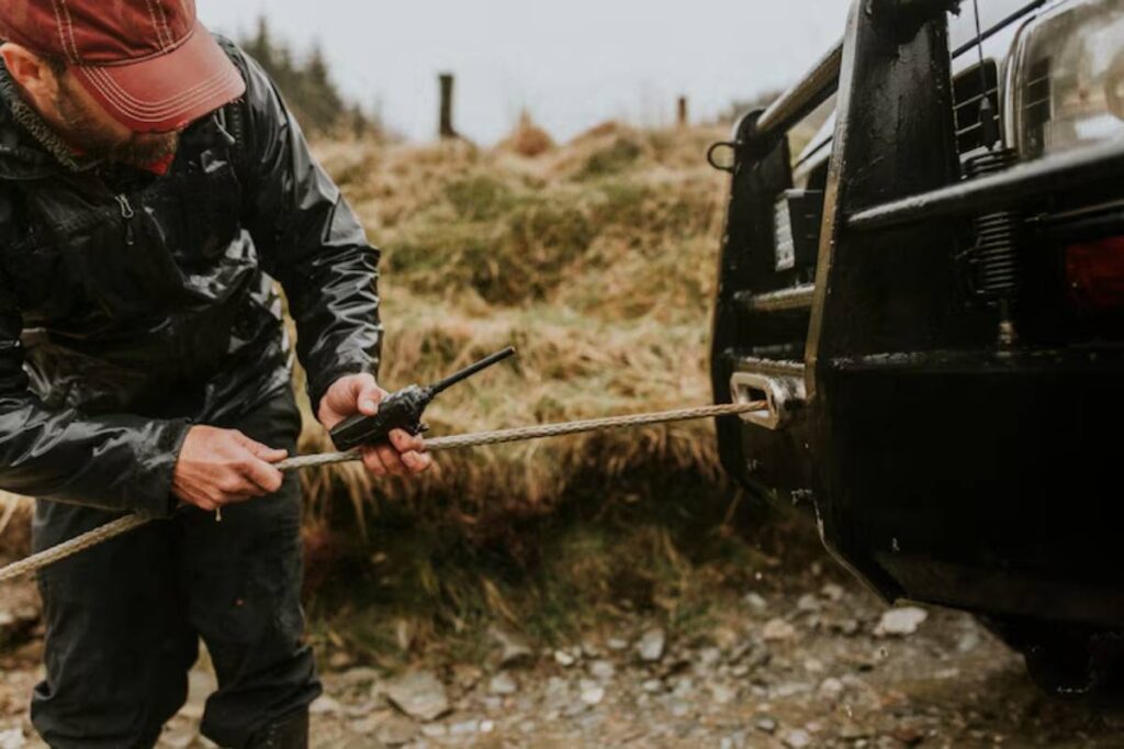A man in a black rain jacket uses a walkie-talkie while securing a rope to a winch on a rugged vehicle in a grassy setting.