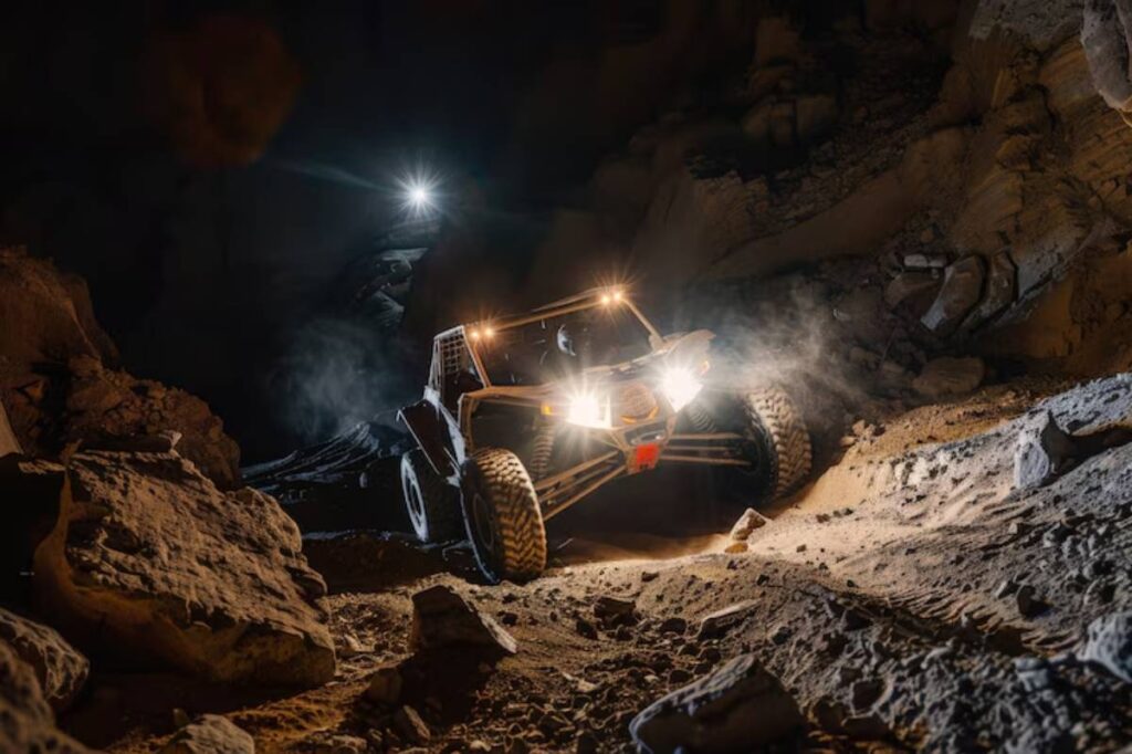 A rugged off-road vehicle navigates a rocky, dimly lit cave, illuminated by bright headlights and dust swirling around.