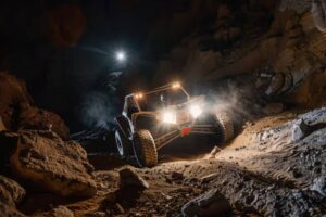 A rugged off-road vehicle navigates a rocky, dimly lit cave, illuminated by bright headlights and dust swirling around.