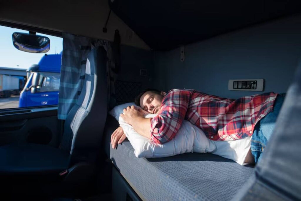 A person rests on a pillow inside a truck cabin, with sunlight streaming in through the window and blue trucks visible outside.