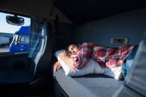 A person rests on a pillow inside a truck cabin, with sunlight streaming in through the window and blue trucks visible outside.