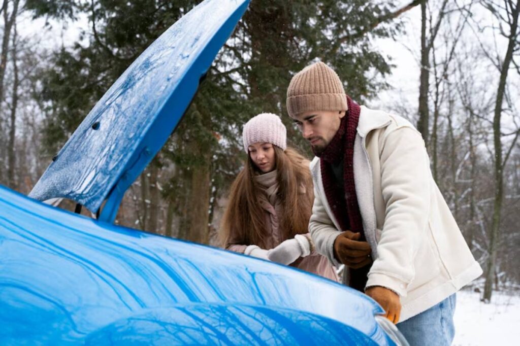 A man and a woman inspect the engine of a blue car in a snowy forest, both dressed warmly for winter.