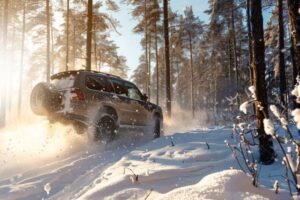A rugged SUV navigating through a snow-covered forest, spraying snow and dirt, with sunlight filtering through tall trees.