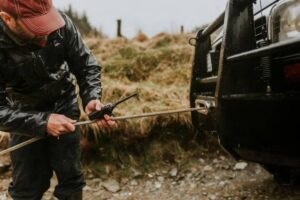 A man in a black rain jacket uses a walkie-talkie while securing a rope to a winch on a rugged vehicle in a grassy setting.