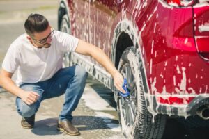 A person kneels beside a shiny red car, scrubbing its wheel with a blue cloth while surrounded by soap suds.