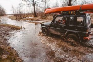 A rugged black SUV drives through a muddy waterlogged road, with a bright orange kayak secured on its roof.