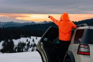 A person in an orange jacket stands by a car, pointing towards a sunset over snow-covered mountains and pine forests.