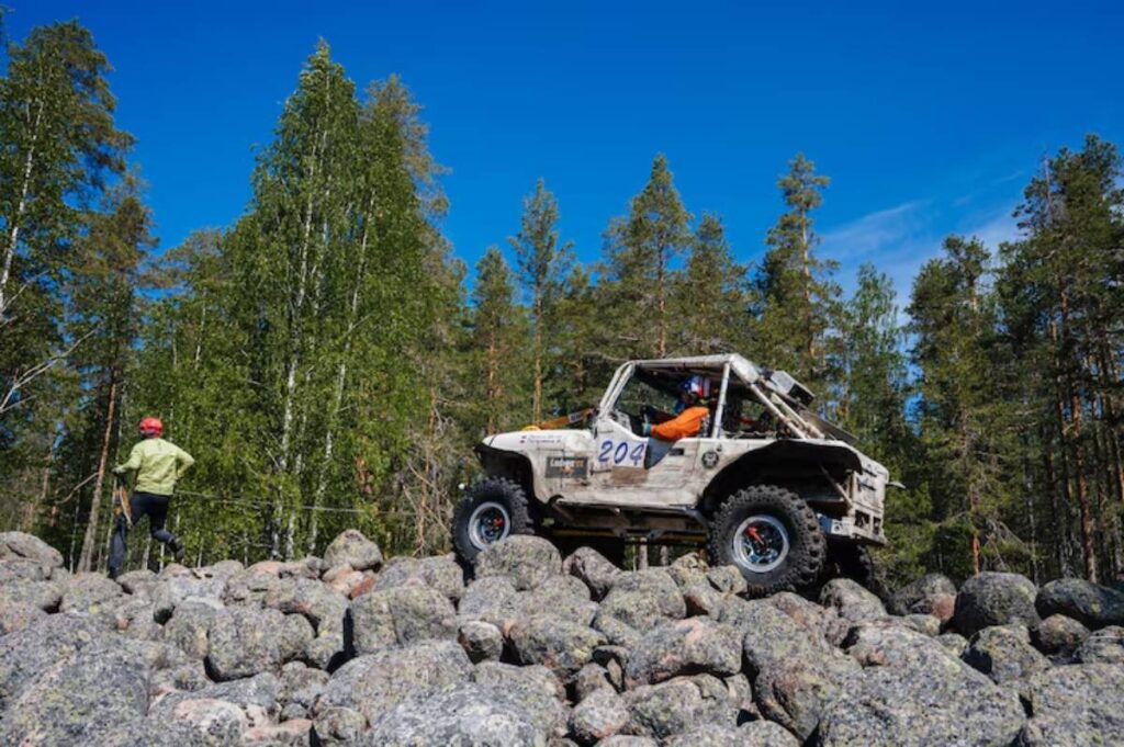 A rugged off-road vehicle navigates a rocky terrain under a clear blue sky, with a person in a helmet observing nearby.