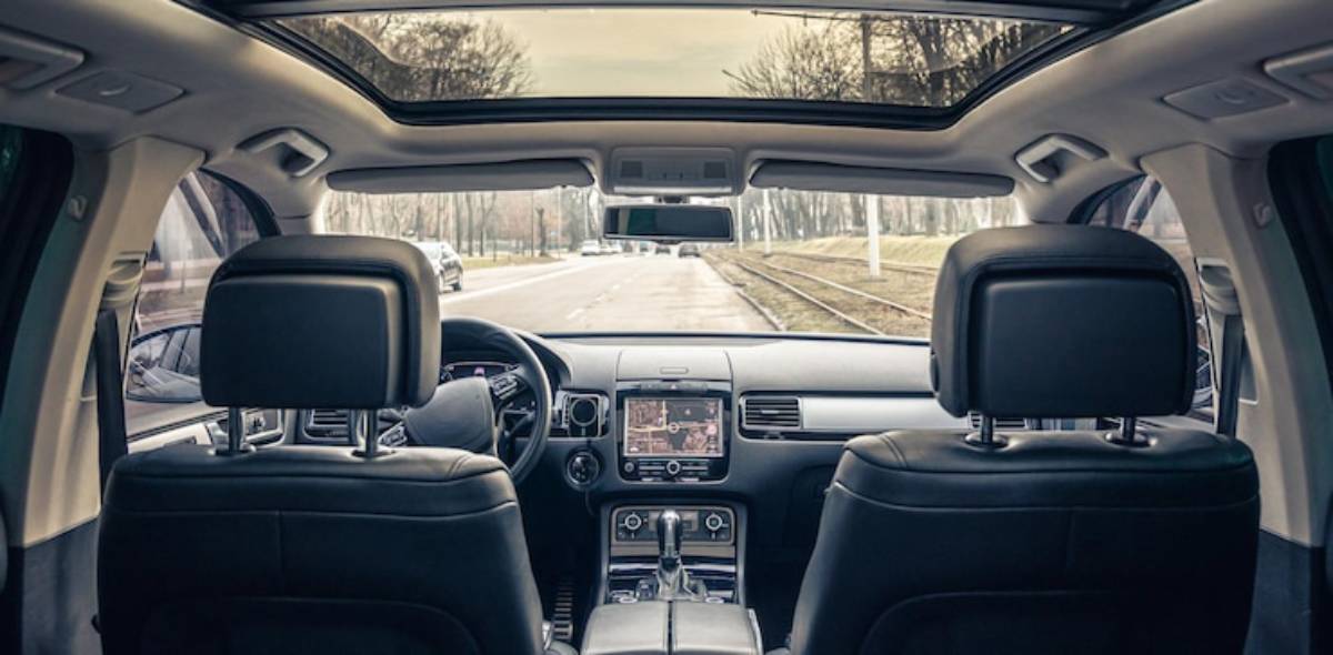 View from the backseat of a luxury car, showing the driver, dashboard, and a sunroof, with a tree-lined street outside.