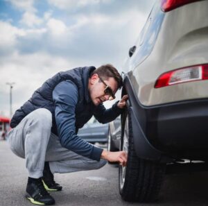 A person crouches beside a parked car, inspecting the tire, wearing a dark vest and sweatpants in a busy parking lot.